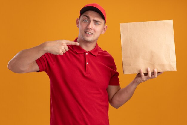 Pleased young delivery man wearing uniform with cap holding and points at paper food package isolated on orange wall