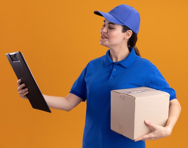 Pleased young delivery woman in uniform and cap holding cardbox and clipboard looking at clipboard 