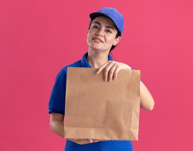 Довольная молодая женщина-доставщик в униформе и кепке, держащая бумажный пакет на розовой стене с копией пространства