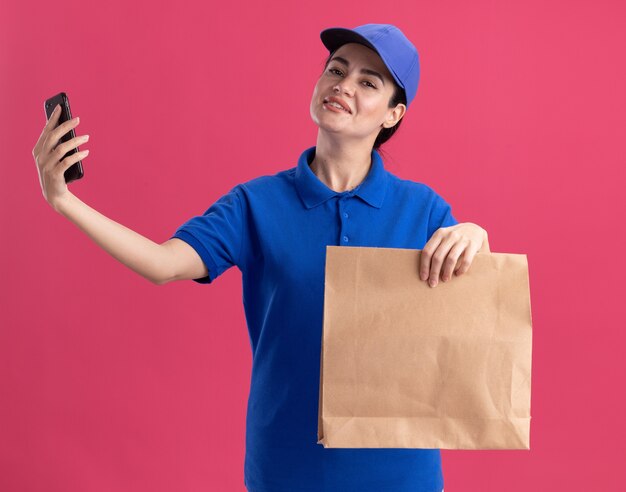 Pleased young delivery woman in uniform and cap holding paper package and mobile phone 