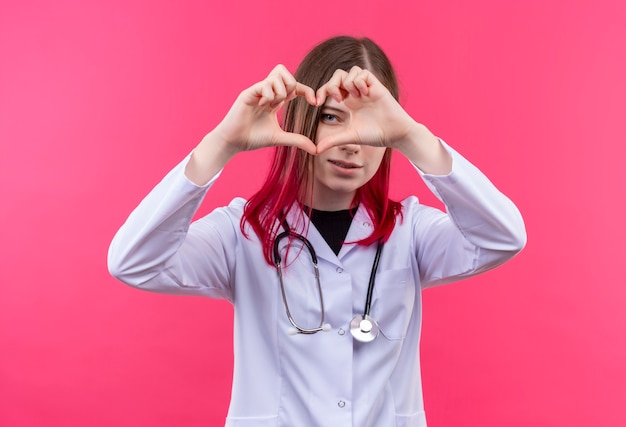 ピンクの孤立した背景に心臓のジェスチャーを示す聴診器の医療用ガウンを身に着けている若い医者の女の子を喜ばせる