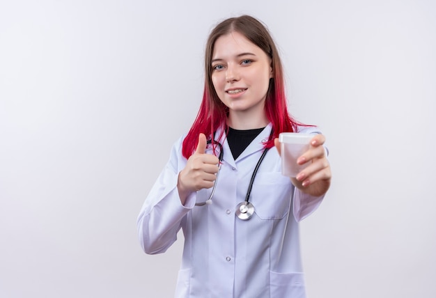 Pleased young doctor girl wearing stethoscope medical robe holding empty can her thumb up on isolated white background