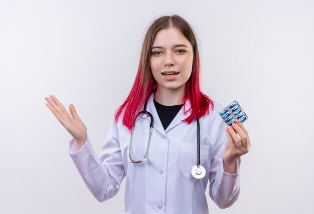 Pleased young doctor girl wearing stethoscope medical robe holding pills spread hand on isolated white background