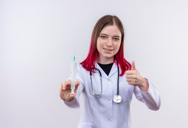 Pleased young doctor girl wearing stethoscope medical robe holding syringe her thumb up on isolated white background