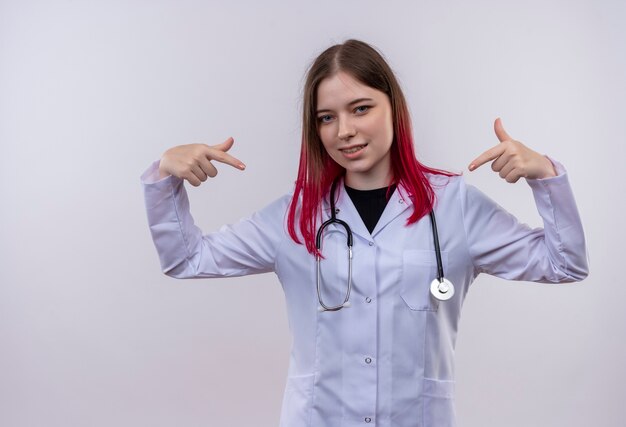 Довольная молодая девушка-врач в медицинском халате со стетоскопом указывает на себя на изолированном белом фоне