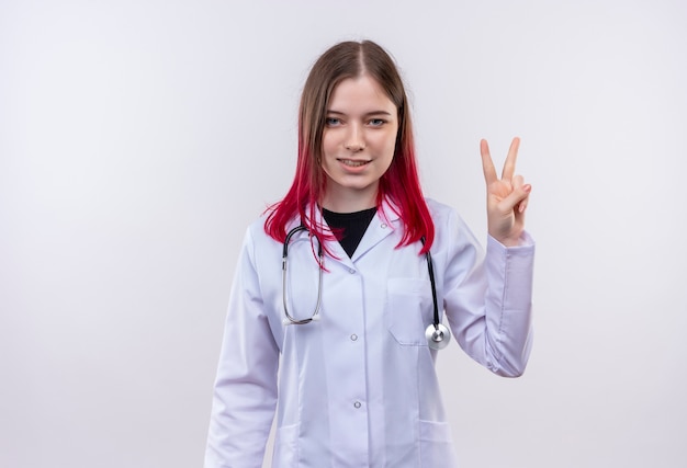 격리 된 흰색 배경에 평화 제스처를 보여주는 청진 의료 가운을 입고 기쁘게 젊은 의사 소녀