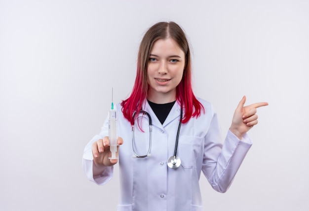 孤立した白い壁に注射器を持って聴診器の医療ローブを身に着けている若い医者の女性が指を横に向けて喜んで