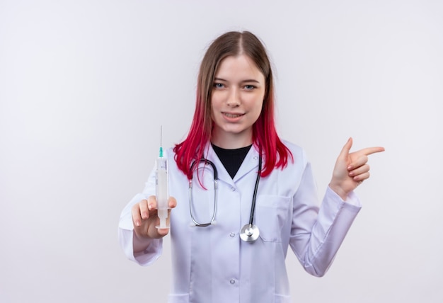  pleased young doctor woman wearing stethoscope medical robe holding syringe points finger to side on isolated white wall