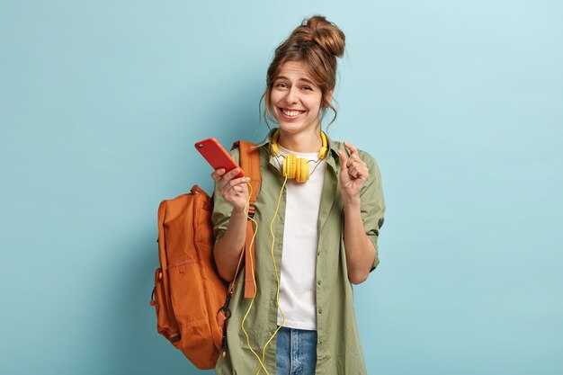 Pleased young European girl shows something very little with hand, enjoys free time and surfes internet on mobile phone