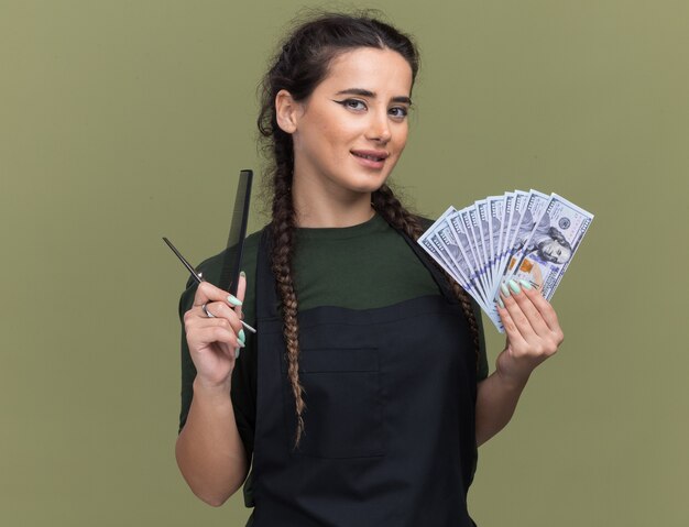 Lieta giovane donna barbiere in uniforme che tiene contanti con strumenti di barbiere isolati sulla parete verde oliva