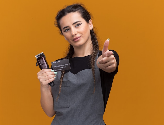 Pleased young female barber in uniform holding credit card and hair clippers points  isolated on orange wall