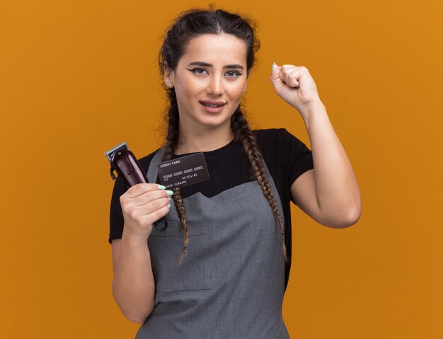 Довольная молодая женщина-парикмахер в униформе держит кредитную карту и машинку для стрижки волос, показывая жест да, изолированный на оранжевой стене