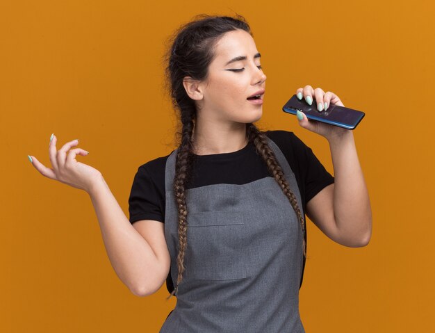 Pleased young female barber in uniform holding phone and singing isolated on orange wall