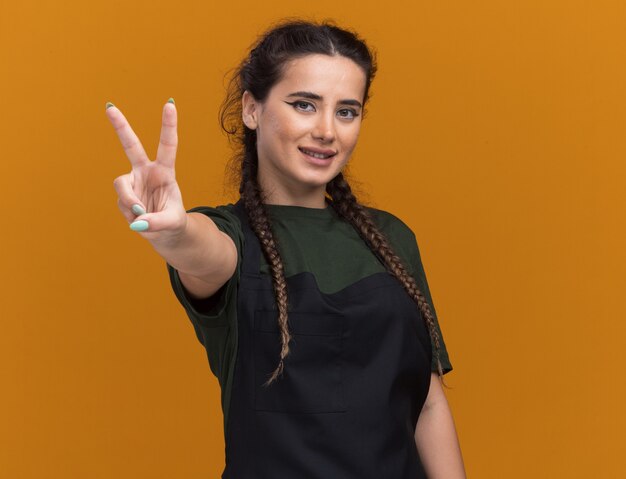 Довольная молодая женщина-парикмахер в униформе, демонстрирующая жест мира на оранжевой стене с копией пространства