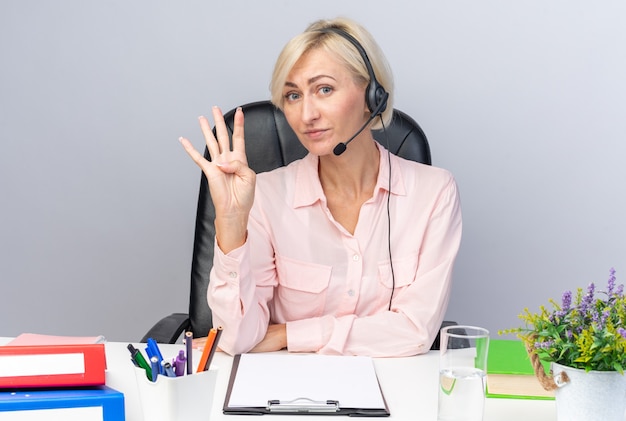 Бесплатное фото Довольная молодая женщина-оператор call-центра в гарнитуре сидит за столом с офисными инструментами, показывающими четыре