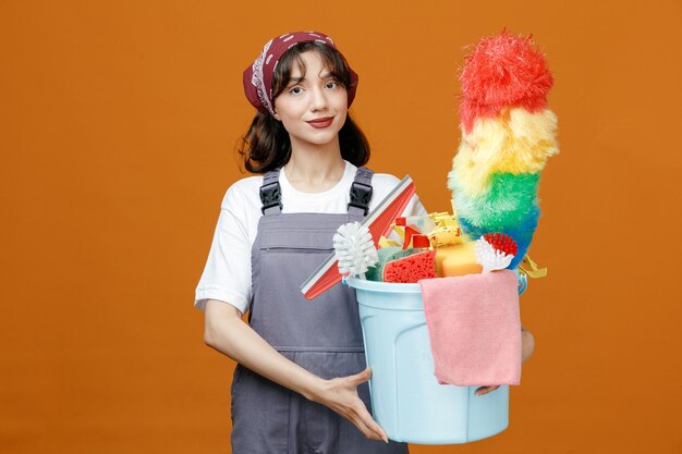 주황색 배경에 격리된 카메라를 바라보는 청소 도구가 있는 양동이를 들고 제복을 입고 반다나를 입은 젊은 여성 청소부