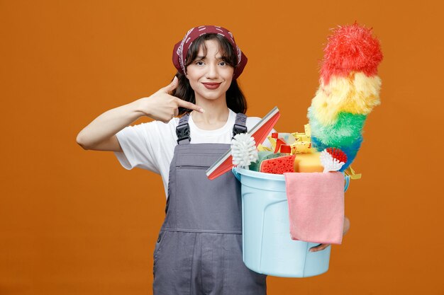 Pleased young female cleaner wearing uniform and bandana holding and pointing at bucket of cleaning tools looking at camera isolated on orange background