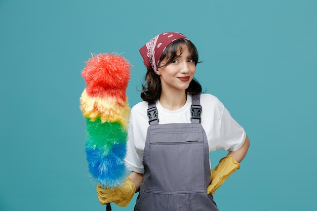 Free photo pleased young female cleaner wearing uniform bandana and rubber gloves holding feather duster looking at camera while keeping hand on waist isolated on blue background