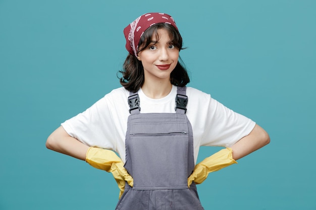 Felice giovane donna addetta alle pulizie che indossa bandana uniforme e guanti di gomma che tengono le mani sulla vita guardando la fotocamera isolata su sfondo blu