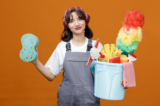 Pleased young female cleaner wearing uniform and bandana showing sponge holding bucket of cleaning tools looking at camera isolated on orange background