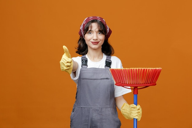 Pleased young female cleaner wearing uniform rubber gloves and bandana holding squeegee mop looking at camera stretching hand out showing thumb up isolated on orange background