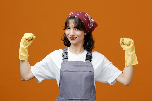 オレンジ色の背景に分離された強いジェスチャーを作るカメラを見て均一なゴム手袋とバンダナを身に着けている若い女性クリーナーを喜ばせる