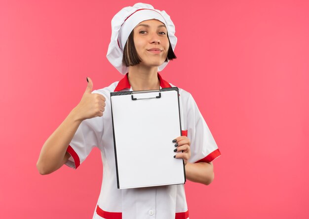 Pleased young female cook in chef uniform holding clipboard and showing thumb up isolated on pink background with copy space