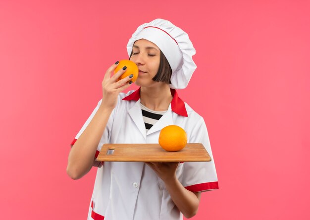 その上にオレンジ色のまな板を保持し、ピンクの壁に隔離された目を閉じてオレンジ色を嗅ぐシェフの制服を着た若い女性料理人を喜ばせる