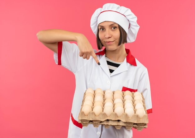 Free photo pleased young female cook in chef uniform holding and pointing at carton of eggs isolated on pink background