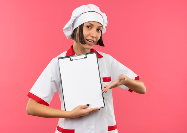 Pleased young female cook in chef uniform holding and pointing with hand at clipboard isolated on pink background