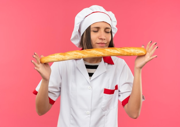 Довольная молодая женщина-повар в униформе шеф-повара держит и нюхает хлебную палочку с закрытыми глазами, изолированными на розовом фоне