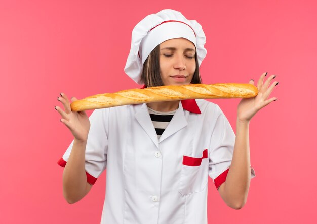 ピンクの背景で隔離の目を閉じてパン棒を保持し、嗅ぐシェフの制服を着た若い女性料理人を喜ばせる