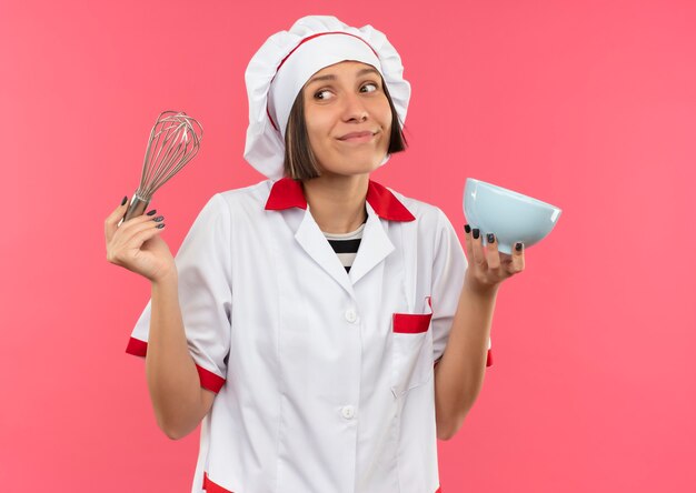 Pleased young female cook in chef uniform holding whisk and bowl and looking at side isolated on pink wall