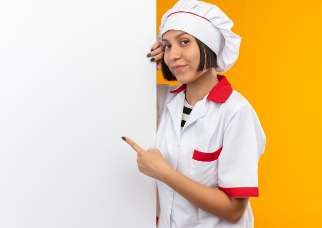 뒤에 서서 벽에 고립 된 흰 벽을 가리키는 요리사 유니폼에 기쁘게 젊은 여성 요리사