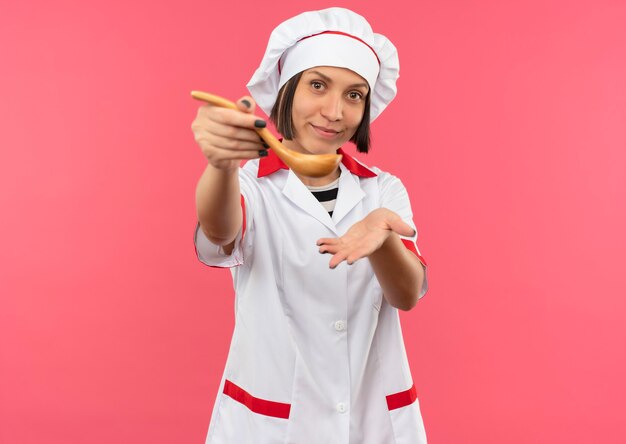 Pleased young female cook in chef uniform stretching out spoon towards front and showing empty hand isolated on pink wall