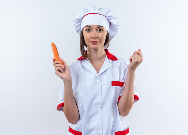 Pleased young female cook wearing chef uniform holding carrot isolated on white background