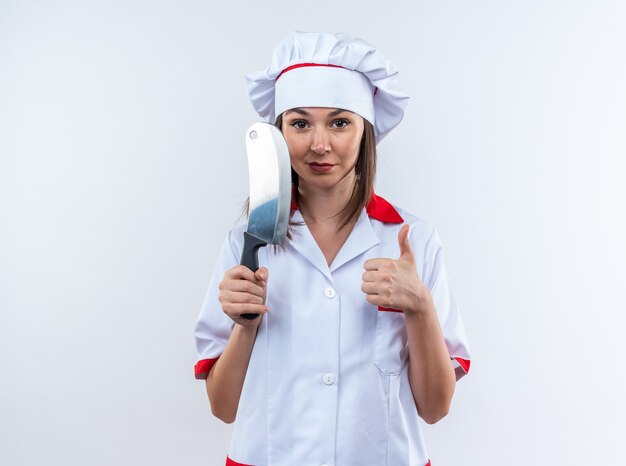 흰색 벽에 고립 된 엄지 손가락을 보여주는 식칼을 들고 요리사 유니폼을 입고 기쁘게 젊은 여성 요리사