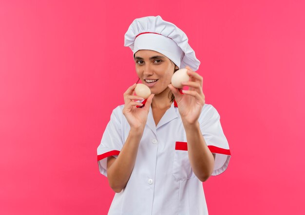 Pleased young female cook wearing chef uniform holding eggs on isolated pink wall with copy space