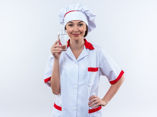 Pleased young female cook wearing chef uniform holding glass of water putting hand on hip isolated on white background