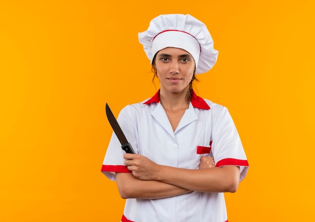 Довольная молодая женщина-повар в униформе шеф-повара держит нож и скрещивает руки на изолированной желтой стене с копией пространства