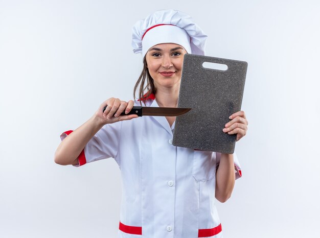 pleased young female cook wearing chef uniform holding knife with cutting board isolated on white wall