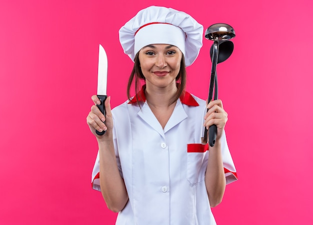 Довольная молодая женщина-повар в униформе шеф-повара держит нож с ковшом на розовом фоне