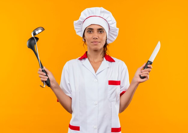 복사 공간이 격리 된 노란색 벽에 국자와 칼을 들고 요리사 유니폼을 입고 기쁘게 젊은 여성 요리사