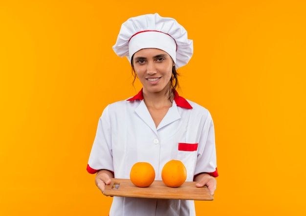 Pleased young female cook wearing chef uniform holding orange on cutting board on isolated yellow wall with copy space