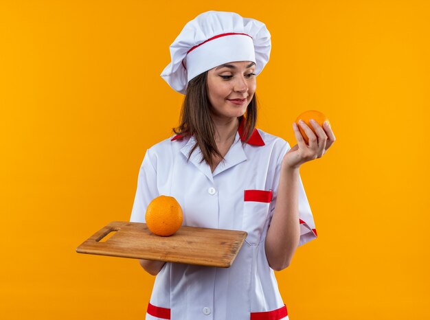 Довольная молодая женщина-повар в униформе шеф-повара держит апельсины на разделочной доске, изолированной на оранжевой стене