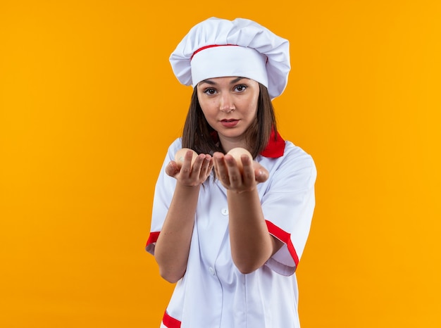 Pleased young female cook wearing chef uniform holding out eggs at camera isolated on orange background