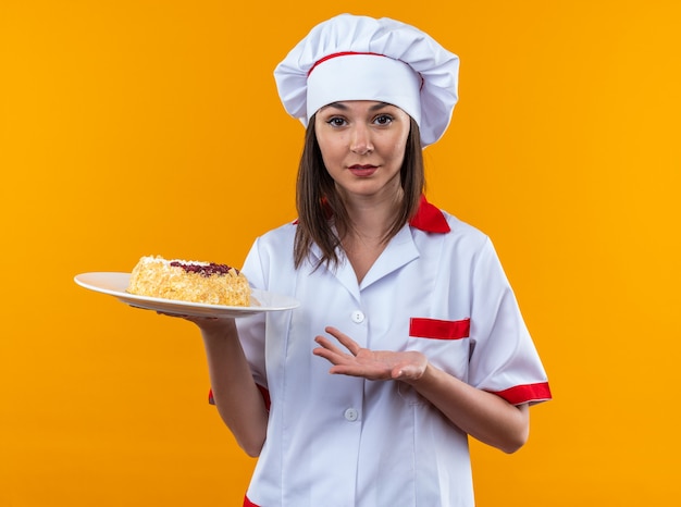 Pleased young female cook wearing chef uniform holding and points with hand at cake on plate isolated on orange background