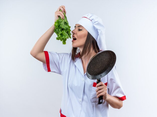 pleased young female cook wearing chef uniform holding salad with frying pan isolated on white wall