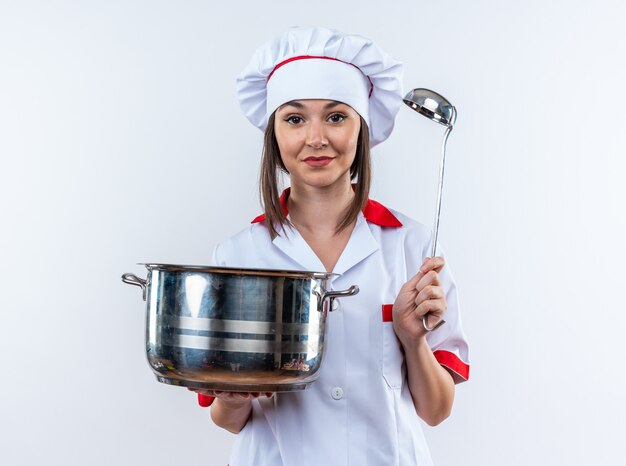 Pleased young female cook wearing chef uniform holding saucepan with ladle isolated on white background