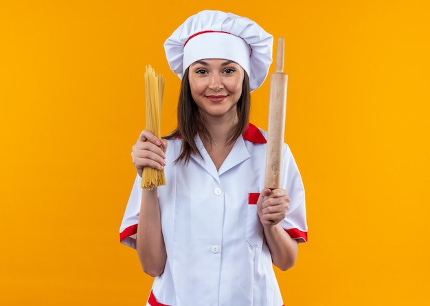 오렌지 배경에 고립 된 롤링 핀 스파게티를 들고 요리사 유니폼을 입고 기쁘게 젊은 여성 요리사